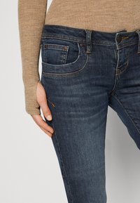 Vaqueros de mezclilla azul oscuro con una textura envejecida, con costuras naranjas contrastantes y un bolsillo delantero. Un top ribeteado beige es parcialmente visible.