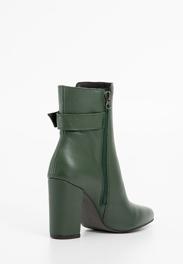 Zielona skórzana botka sięgająca kostki z zapięciem na zamek, kwadratowym noskiem i masywnym obcasem. Posiada gładkie wykończenie oraz regulowany detal w formie paska.