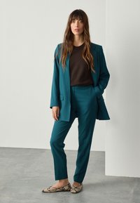 Teal suit made of smooth fabric, featuring a tailored blazer and slim-fit trousers. Paired with a brown top and snake-pattern flat shoes.