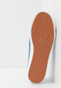Semelle de chaussure avec grip en caoutchouc marron texturé reposant verticalement contre un angle blanc de mur et de sol.