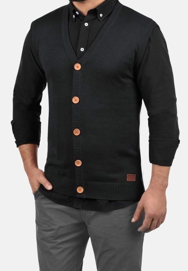 Homme portant un cardigan noir boutonné sur une chemise noire aux manches retroussées et un pantalon gris, debout avec une main dans la poche.