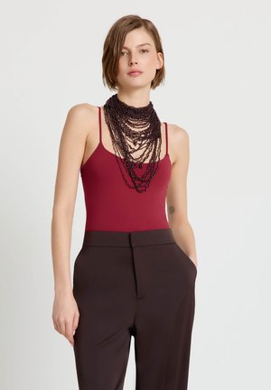 Donna con top bordeaux senza maniche, collana a strati con perline scure e pantaloni marrone scuro a vita alta, con le mani in tasca.