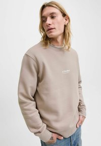 Beež sweatshirt, millel on ümar kaelus ja valge tikkimine tekstiga "JJ Studios". Mudel kannab kulunud siniseid teksaseid.