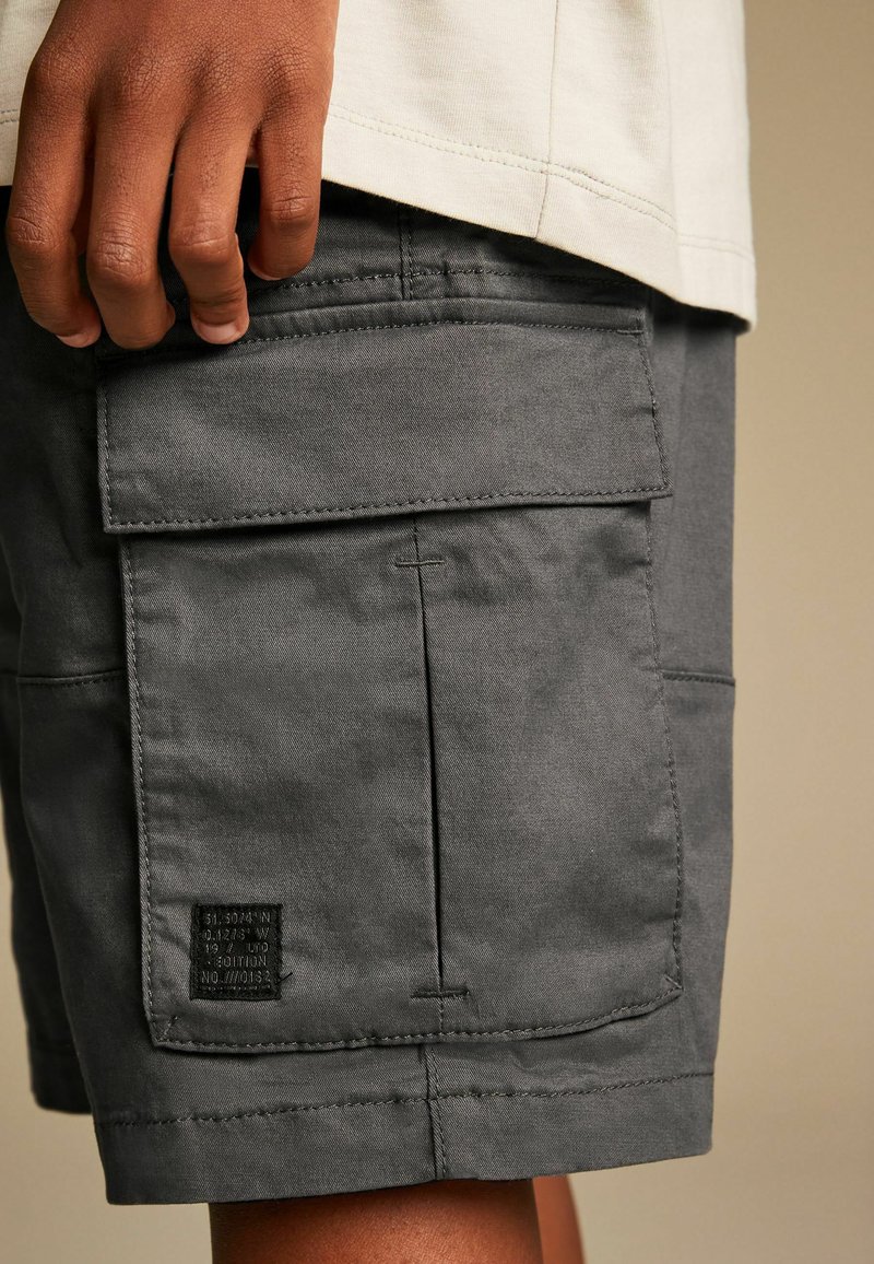 Bermudas grises avec plusieurs poches, un design cousu et un tissu texturé. Comprend une étiquette de marque sur la poche arrière.