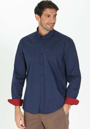 Chemise bleu marine à manches longues avec des poignets rouges, dotée d'un col, d'une coupe ajustée et de boutons contrastants. Texture du tissu lisse.