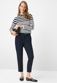 Marineblauwe taps toelopende broek met voorzakken, gecombineerd met een zwart-wit gestreept shirt met lange mouwen en zwarte mesh ballerina's, met een zwarte handtas in de hand.