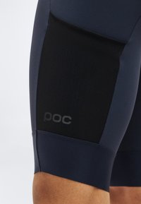 Κοντινό πλάνο στο μηρό ενός ατόμου που φοράει μαύρο και σκούρο μπλε ποδηλατικό σορτς με μαύρο πλέγμα και λογότυπο "poc".