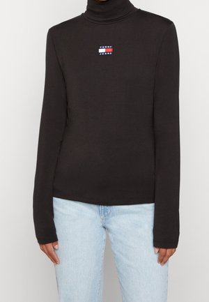 Henkilö, joka pukeutunut mustaan pitkähihainen poolopaitaan, jossa Tommy Jeans -logo rinnassa, ja vaaleansiniset farkut, seisoo yksinkertaisen taustan edessä.