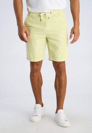 Uomo con pantaloncini di velluto a coste giallo chiaro con coulisse e sneakers bianche in piedi davanti a uno sfondo semplice.
