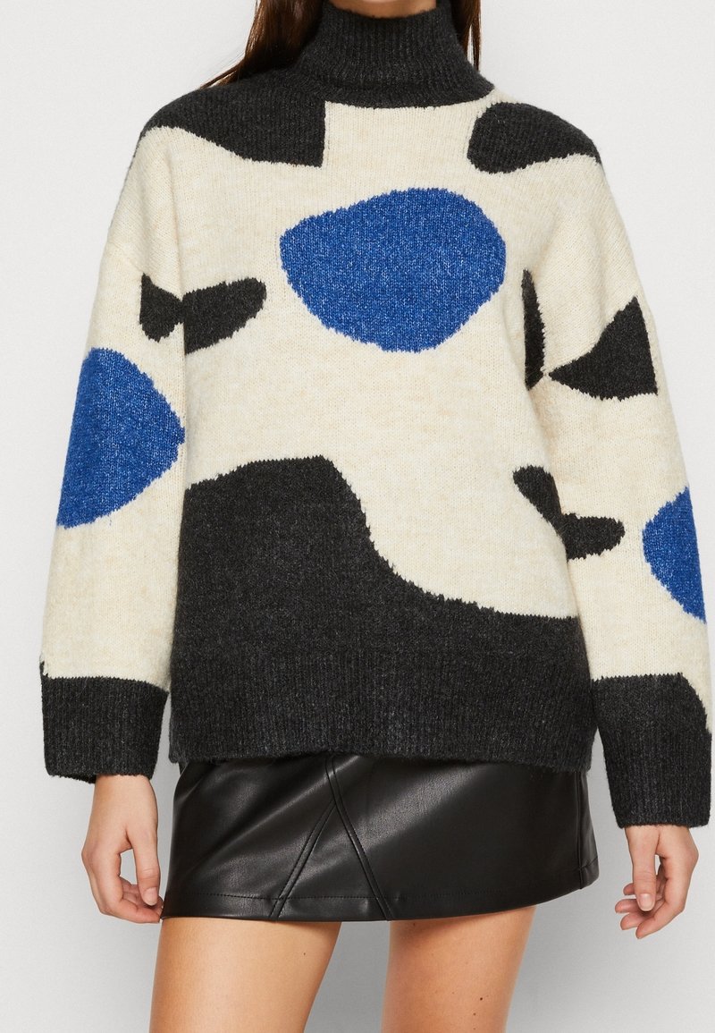 Femme portant un pull à col roulé à motif abstrait noir, blanc et bleu avec une mini-jupe en cuir noire.
