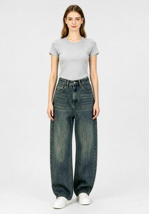 Giovane donna che indossa una t-shirt grigio chiaro, jeans blu sbiaditi a vita alta e a gamba larga e sneakers bianche, in piedi davanti a uno sfondo semplice.