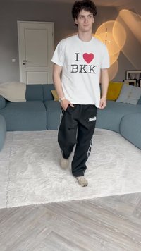 Bela majica s natpisom "I ❤️ BKK", črne trenirke s stranskimi črtami, hodi po svetlo obarvanem puhastem preprogo v opremljeni sobi.
