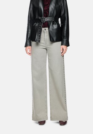 Person wearing light gray wide-leg pants with side studs, a black belted leather jacket, patterned dark red top, and dark shoes.