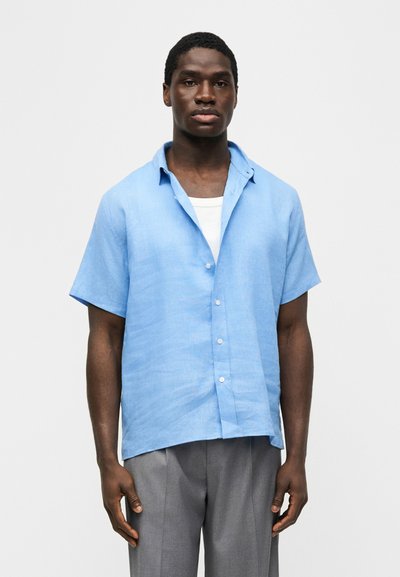 Άνδρας που φοράει ανοιχτό μπλε πουκάμισο με κοντά μανίκια πάνω από λευκό εσώρουχο και γκρι παντελόνι, στέκεται μπροστά σε απλό φωτεινό φόντο.
