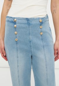 Γαλάζια τζιν παντελόνια ψηλόμεσα με δύο κάθετες σειρές από τέσσερα διακοσμητικά χρυσά κουμπιά μπροστά, φορεμένα με λευκό τοπ.