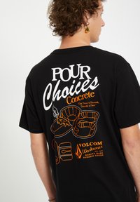 T-shirt en coton noir avec des textes et illustrations graphiques blancs et oranges au dos, incluant "Pour Choices" et un logo.