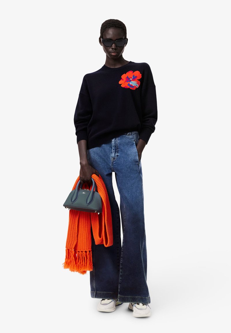 Marineblauer Pullover mit orangefarbenem Blumenausstickerei, kombiniert mit ausgestellten Jeans. Hält eine kleine dunkelgrüne Handtasche und einen orangefarbenen Schal.