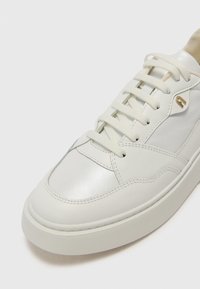 Sneaker en cuir blanc avec une texture lisse, comportant six œillets, des coutures ton sur ton et un accent métallique près des lacets.