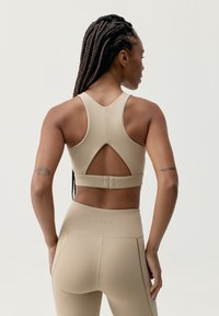 Béžový sportovní podprsenka s designem racerback, obsahující vyřezávaný detail a bezpečné zapínání na zádech. Materiál je hladký a elastický.