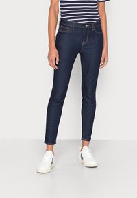 Jeans slim fit de color azul oscuro hechos de mezclilla, con costuras naranjas en contraste, cinco bolsillos y cierre de cremallera, combinados con zapatillas blancas.