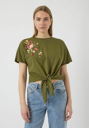 T-shirt olive, à manches courtes, avec broderie florale sur le côté gauche et un détail de nœud à l'ourlet. Associé avec un jean bleu.