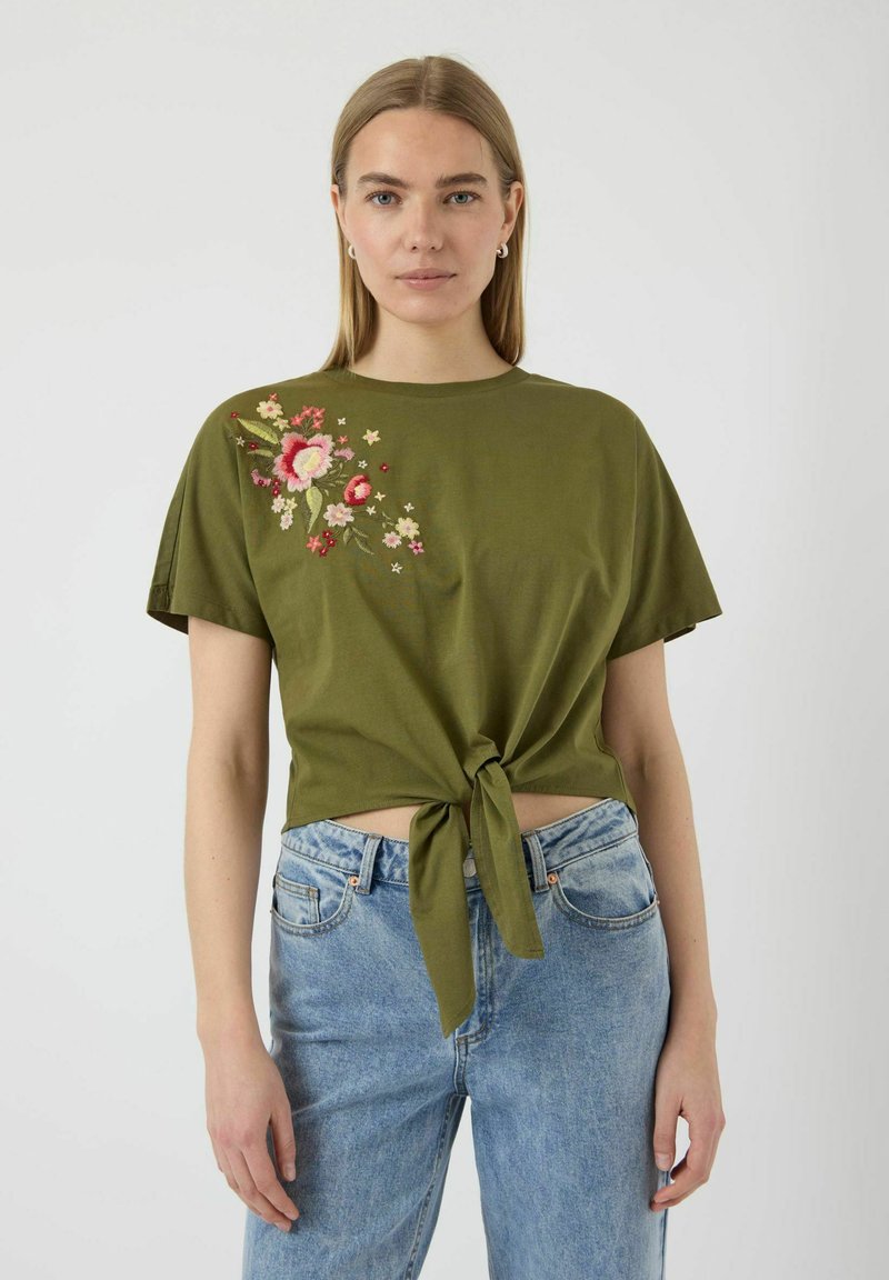T-shirt verde oliva a maniche corte con ricami floreali sul lato sinistro e un dettaglio con nodi sull'orlo anteriore. Abbinato a jeans blu.