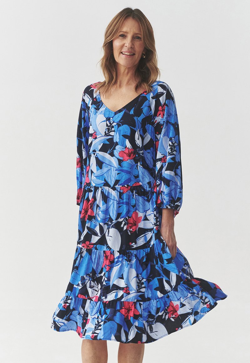 Vestido floral con colores azul, rojo y negro, que presenta un escote en V, mangas largas, volantes en capas y una tela ligera y fluida.