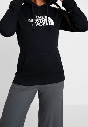 Osoba nosząca czarną bluzę z kapturem The North Face z kieszenią kangurką i szare szerokie spodnie, regulująca sznurki kaptura.