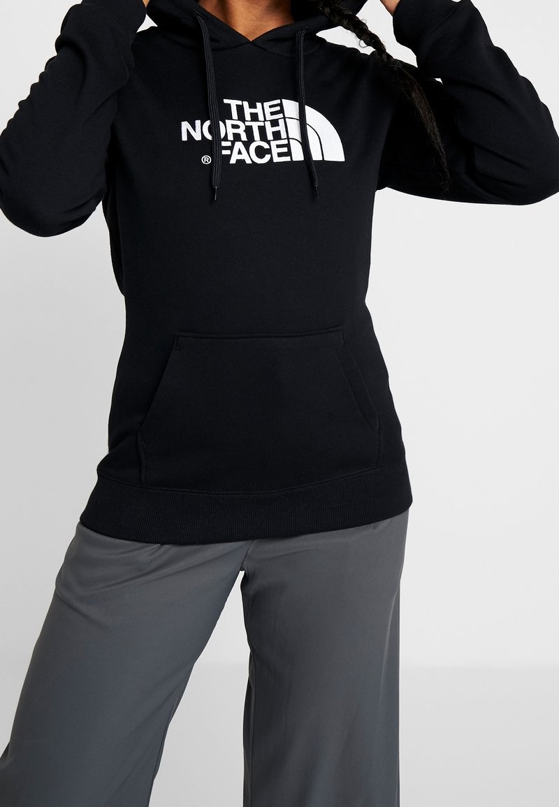 Personne portant un sweat à capuche noir The North Face avec poche kangourou et pantalon gris à jambes larges, ajustant les cordons de la capuche.