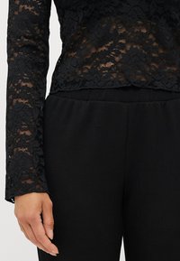 Schwarzes Spitzenoberteil mit langen Ärmeln, floralem Muster und gewelltem Saum, kombiniert mit schwarzen Stoffhosen. Die strukturierte Spitze steht im Kontrast zum glatten Stoff.