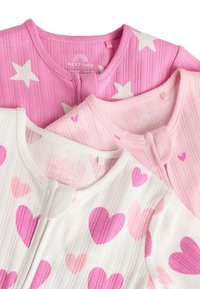 Trois cardigans pour bébés : un rose avec des étoiles blanches, un rose avec de petits cœurs, et un blanc avec de grands cœurs roses. Tous présentent une texture côtelée.