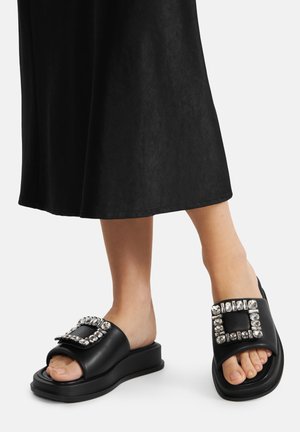 Černé sandály na navlékání s robustní podrážkou, zdobené dekorativní přezkou s kamínky na vršku a hladkou koženou texturou.