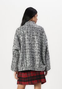 Veste oversized en sequins argentés avec un col montant et des manches longues, associée à une jupe à carreaux rouges. Présente une surface brillante et texturée.
