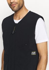Colete preto feito de material leve, com dois bolsos frontais, fecho em zíper e um logótipo com detalhes amarelos no canto inferior direito.