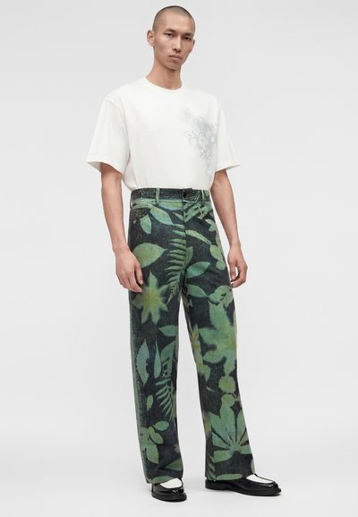 Άνδρας που στέκεται φορώντας λευκό πουκάμισο με κοντά μανίκια και πράσινο φαρδύ παντελόνι με βοτανικό σχέδιο, μαζί με μαύρα και άσπρα loafers.