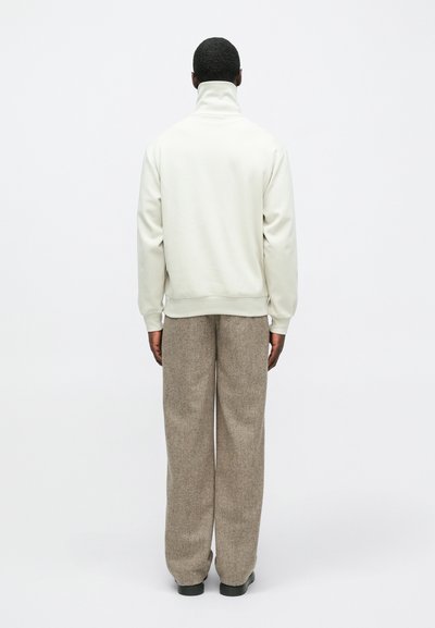 Άτομο που κοιτάζει μακριά, φορώντας ένα λευκό πουλόβερ με ψηλό λαιμό, ανοιχτόχρωμο μπεζ παντελόνι και μαύρα παπούτσια, στέκεται μπροστά σε ουδέτερο λευκό φόντο.