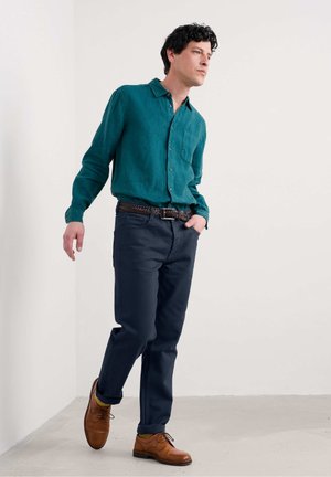 Camisa de lino verde azulado con collar de botón, pantalones oscuros azul marino y zapatos de cuero marrón con un acabado texturizado, acompañados de un cinturón trenzado.