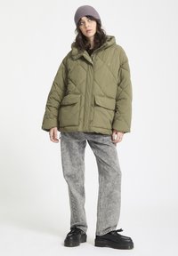 Chaqueta acolchada verde oliva con patrón de rombos, cremallera frontal, dos grandes bolsillos, combinada con jeans grises desgastados y zapatos de plataforma negros.