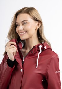 Giacca impermeabile rossa con zip frontale, cappuccio con cordino, dettagli bianchi e polsini elastici. Finitura liscia e lucida con bottoni a scatto.