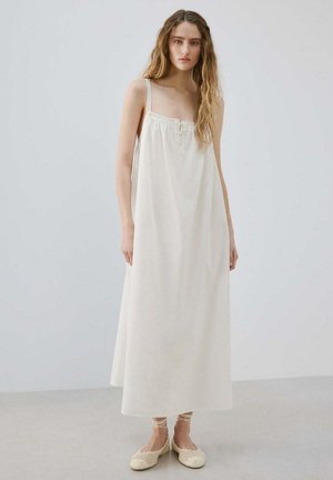 Joven mujer con el pelo largo, vistiendo un vestido blanco suelto y sin mangas y zapatos planos tejidos de color crema con lazos en el tobillo, de pie sobre un suelo gris.