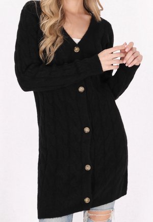 Femme portant un long cardigan noir en maille torsadée avec de gros boutons, associé à un jean bleu clair déchiré, visible du cou jusqu'à la mi-cuisse.