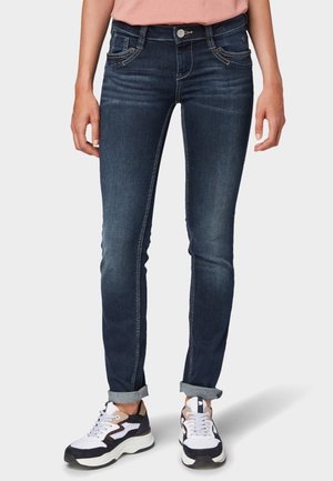 Personne portant un jean slim bleu foncé retroussé et des baskets blanches, noires et beiges, debout les mains détendues le long du corps.