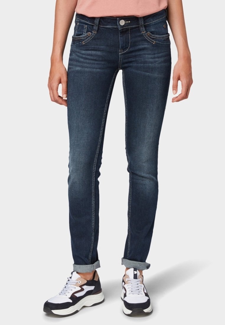Personne portant un jean slim bleu foncé retroussé et des baskets blanches, noires et beiges, debout les mains détendues le long du corps.