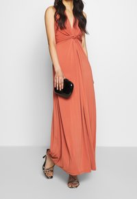 Rostfärgad maxiklänning med knuten front, tillverkad av slät tyg. Kombinerad med en svart clutch och mönstrade klackar med remmar.