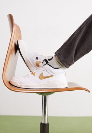 Hvide og guld Nike-sneakers båret af en person med grå bukser, hvilende på en drejelig træstol mod en hvid baggrund.