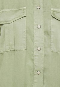 Tissu vert clair avec deux poches à rabat et quatre boutons-pression en métal disposés en une rangée verticale au centre.