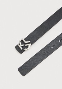 Černý kožený pásek s texturovaným povrchem, který má stříbrnou sponu s logem. Pásek má hladký konec a několik děr pro nastavení.