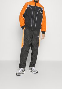 Atletický teplákový set s oranžovou a černou bundou s bílými detaily, doplněný černými kalhotami s oranžovými bočními pruhy a logem.