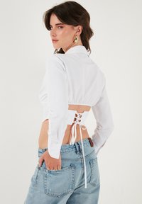 Blusa bianca corta con maniche lunghe e dettaglio di allacciatura sul retro, abbinata a jeans a vita alta di un blu chiaro. Tessuto liscio e leggero.