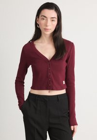 Cardigan scurt de culoare burgundă, cu mâneci lungi, textură ribben și detalii cu butoni pe față. Asortat cu pantaloni negri, cu talie înaltă.